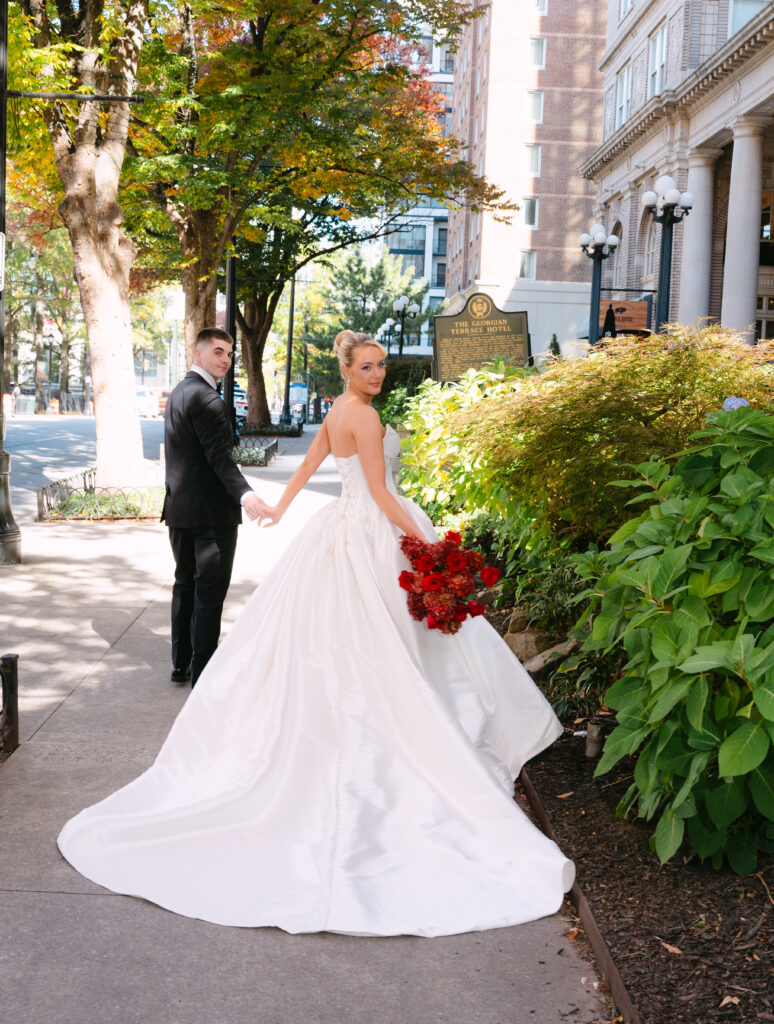 Bride and groom walking hand in hand down the street. The Bride is wearing a silk grown holding red flowers.