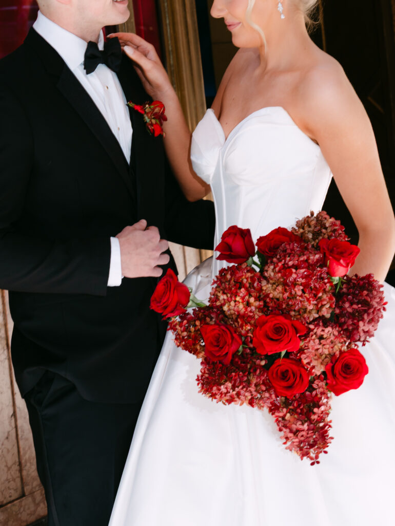 Close-up of the bride and groom standing together outside Fox Theatre. The bride wears a strapless satin gown and holds a bold bouquet of red roses and textured hydrangea, while gently touching the groom’s tuxedo lapel. The groom’s boutonnière matches the deep red florals, creating a romantic Old Hollywood look.