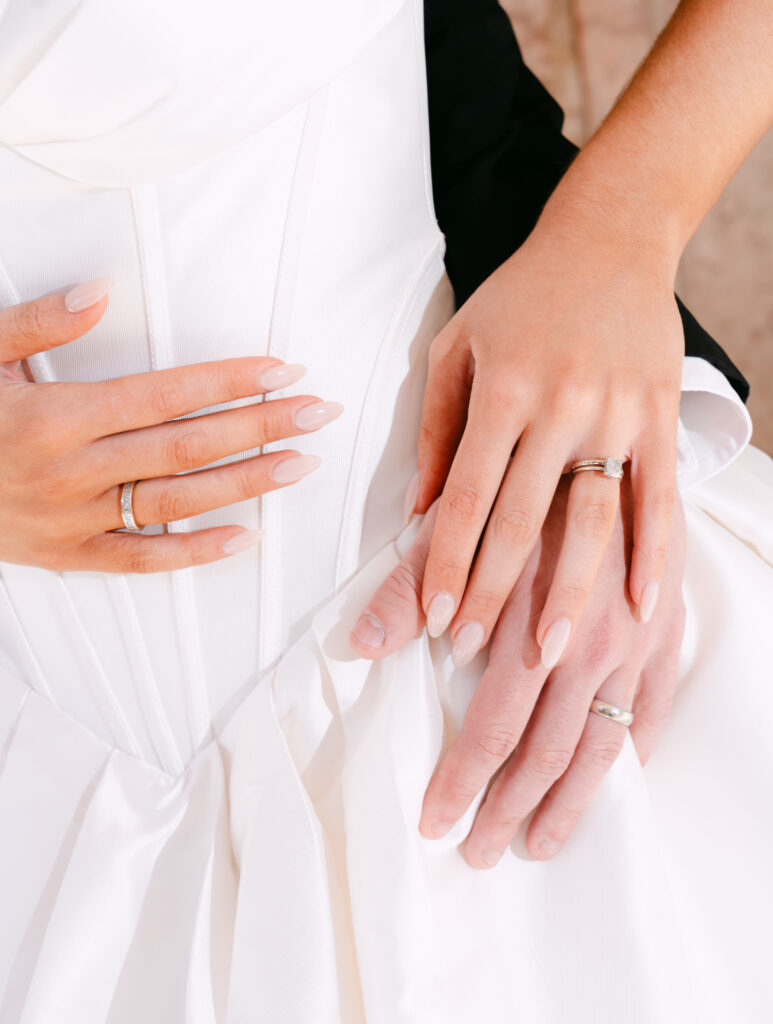 Close-up of the bride and groom’s hands resting on her white satin gown, showcasing their wedding rings and the bride’s nude almond-shaped nails. The groom’s hand gently overlaps hers in a soft, romantic moment.