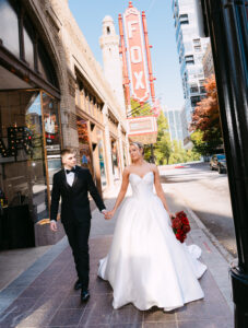 Bride and groom walk hand-in-hand outside the iconic Fox Theatre in Atlanta. The bride wears a glamorous strapless ballgown and carries a bouquet of red roses, while the groom is dressed in a classic black tux. The bright red FOX marquee glows above them as they smile and stroll down the sidewalk.
