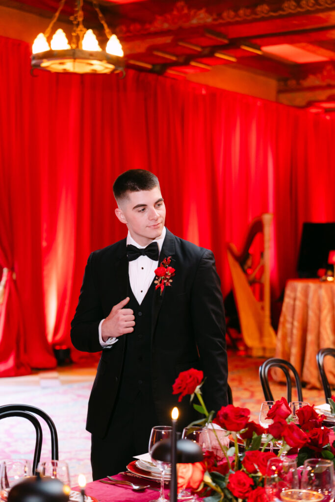 The groom stands in a candlelit reception space draped in rich red fabric at the Fox Theatre in Atlanta. He wears a classic black tux with a red floral boutonnière and looks off to the side with a composed expression. Red rose centerpieces and elegant place settings decorate the foreground, adding to the glamorous, Old Hollywood atmosphere.