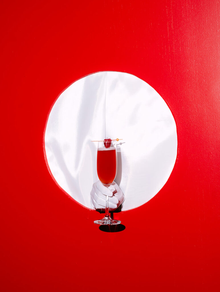 A gloved hand presents a red signature cocktail through a circular white satin cutout in a bold red display wall. The sleek design and dramatic color contrast create a glamorous, modern touch fitting for an Old Hollywood-inspired wedding.