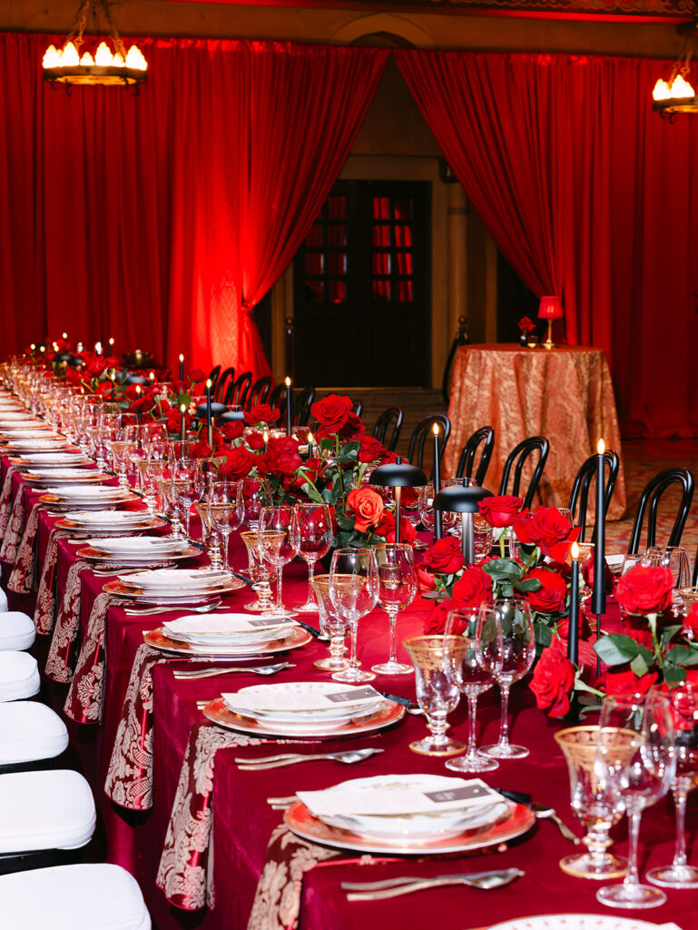 Long banquet tables draped in deep red velvet linens and patterned burgundy runners are set for a wedding reception at the Fox Theatre. The tables feature lush red rose arrangements, black taper candles, gold-rimmed glassware, layered china, and black chairs with white cushions. Dramatic red draping and warm vintage lighting create a glamorous Old Hollywood atmosphere.