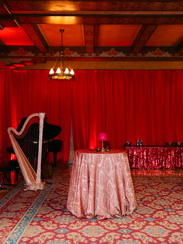 A cocktail table draped in blush patterned linen sits in a richly decorated room with red floor-to-ceiling draping at the Fox Theatre. A harp and grand piano rest in the background, while a glowing red lamp adds a soft touch. The ornate carpet and vintage ceiling details create an elegant, Old Hollywood-inspired atmosphere.