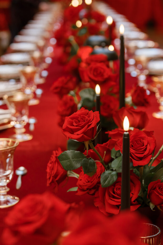 Close-up of a luxurious red rose centerpiece running down a long reception table covered in rich red linens. Black taper candles glow softly among the blooms, surrounded by gold-rimmed glassware and elegant place settings, creating a dramatic Old Hollywood ambiance.