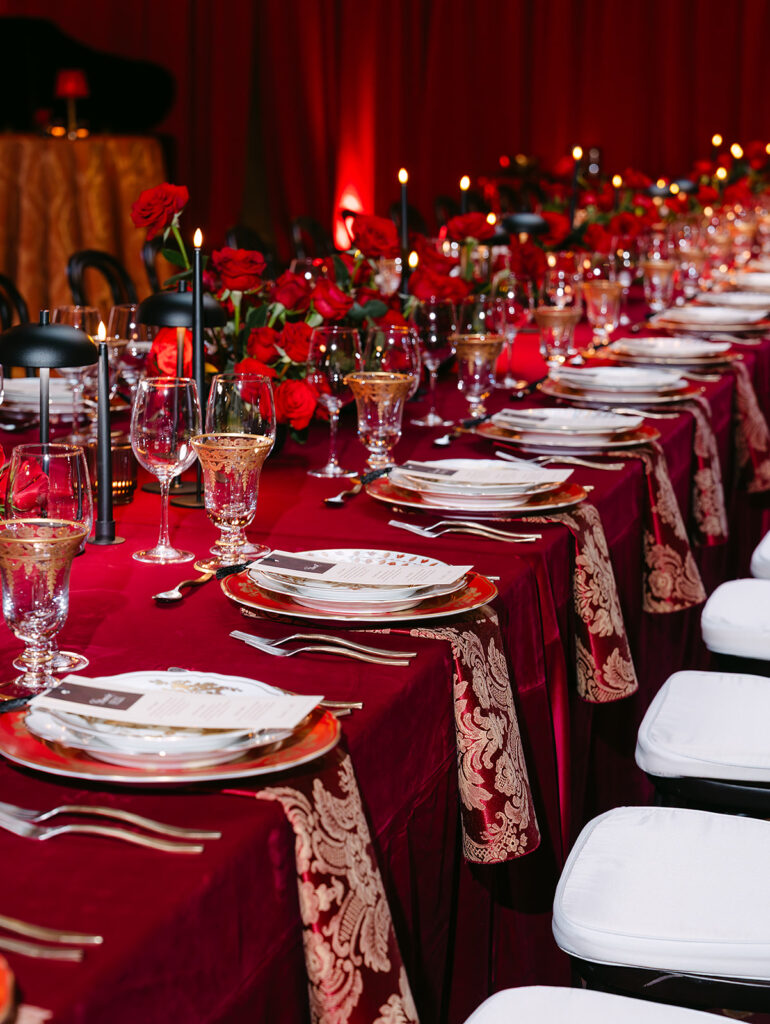 Close-up view of a long wedding reception table draped in deep red velvet linens with ornate burgundy and gold runners. The table is set with layered gold-rimmed china, crystal glassware, polished silverware, and surrounded by white cushioned chairs. Lush red rose arrangements and glowing black taper candles create a dramatic, glamorous Old Hollywood-inspired atmosphere.