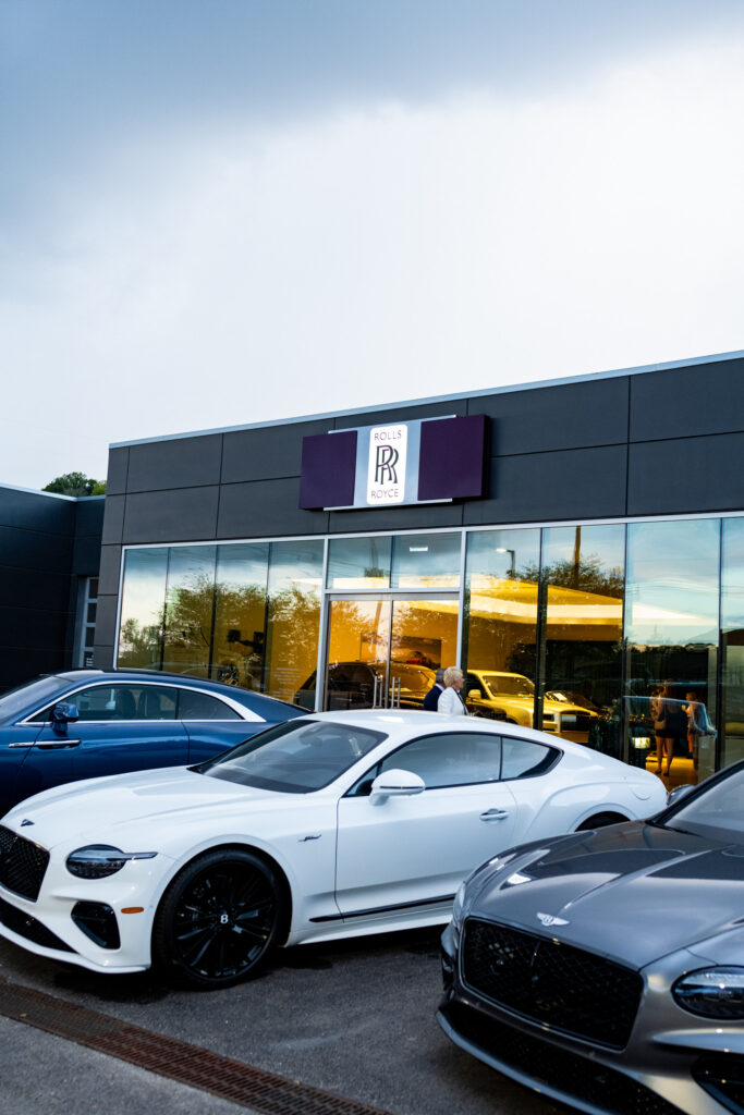 Exterior view of Carlock Motors in Nashville featuring Rolls-Royce branding, with luxury vehicles parked out front during the Modern Luxury Nashville one-year anniversary event, glowing showroom lights visible through glass windows at dusk.