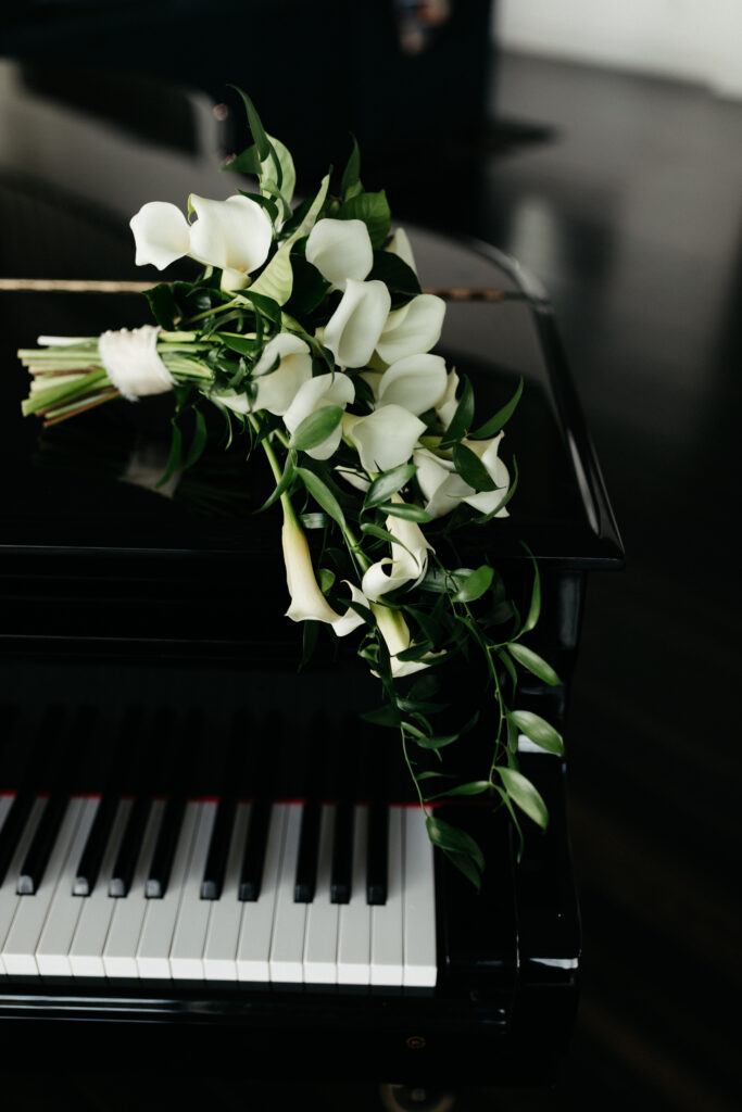 White calla lily bridal bouquet with cascading greenery resting on a black grand piano, styled wedding detail photo