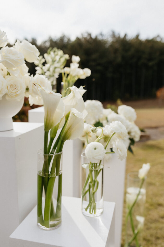 White calla lilies and roses arranged in clear glass cylinders on white pedestals for modern outdoor wedding ceremony décor