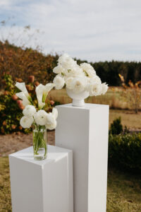 White calla lilies and roses displayed in modern vases on white pedestals for an outdoor wedding ceremony