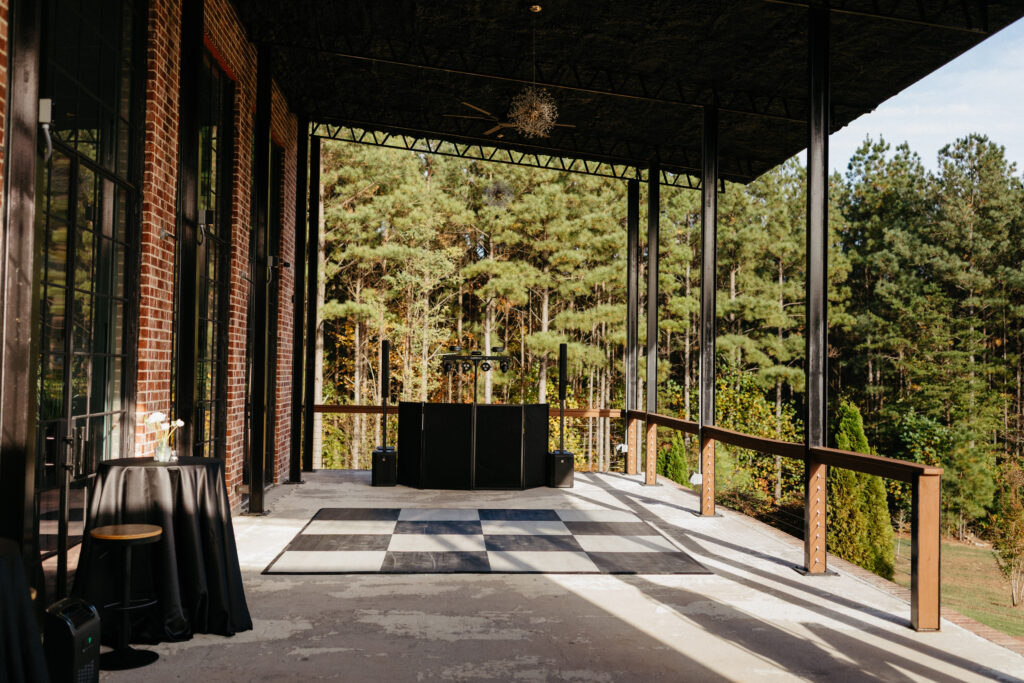 Outdoor wedding reception dance floor on covered terrace with DJ setup, brick walls, and forest views