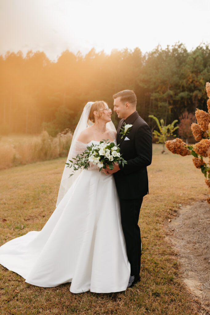 Bride and groom sharing a romantic moment outdoors, bride holding a white calla lily bouquet with greenery at sunset