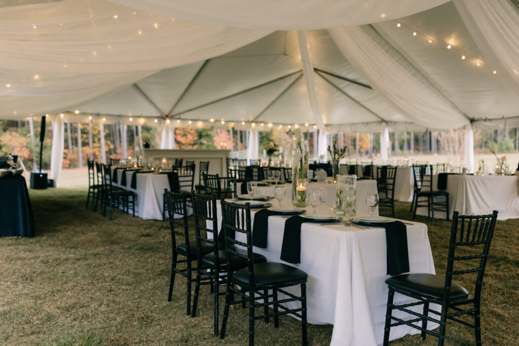 Tented wedding reception with white draping, string lights, long tables, and black chairs styled in an outdoor setting