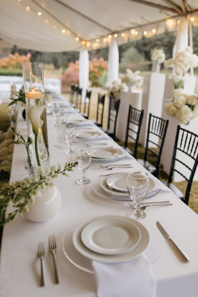 Wedding reception table styled with white linens, calla lily centerpieces, floating candles, and black chairs under a draped tent