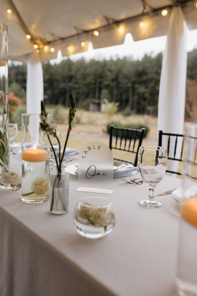 Acrylic wedding table number reading “Table One” styled with candles, glassware, and white floral accents under a draped tent