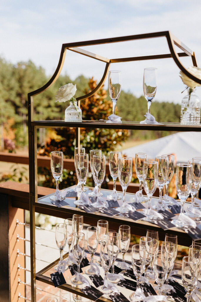 Champagne flutes with escort cards displayed on a gold shelving unit at an outdoor wedding reception overlooking trees